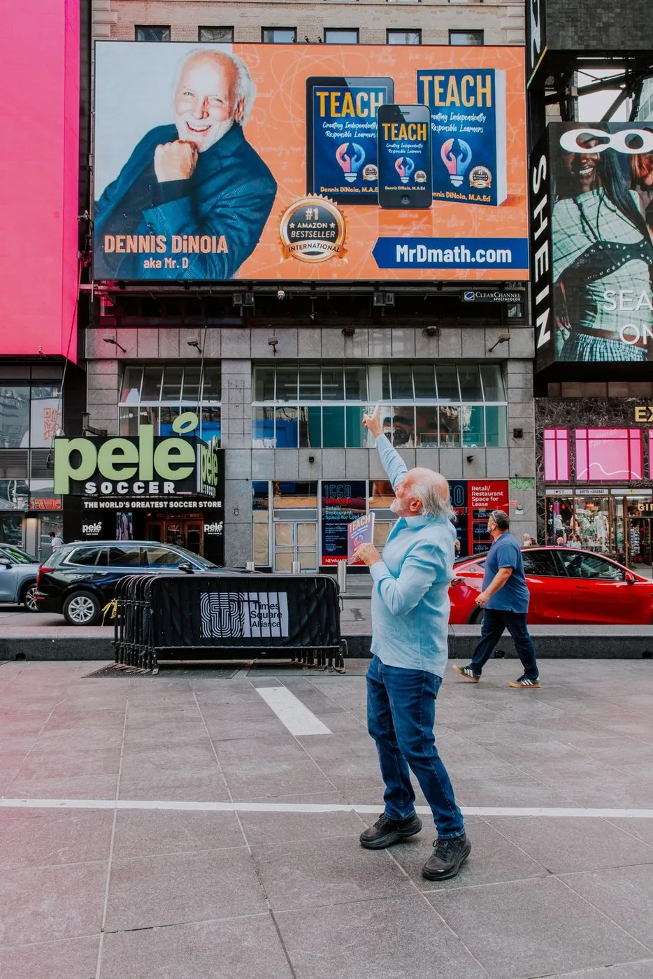 Dennis in Times Square, pointing up at his TEACH billboard, holding the book